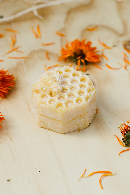 Image of a Busy Bee Soap - a creamy coloured soap moulded to look like honeycomb. Soap on a light wood table surrounded by calendular flowers and petals.